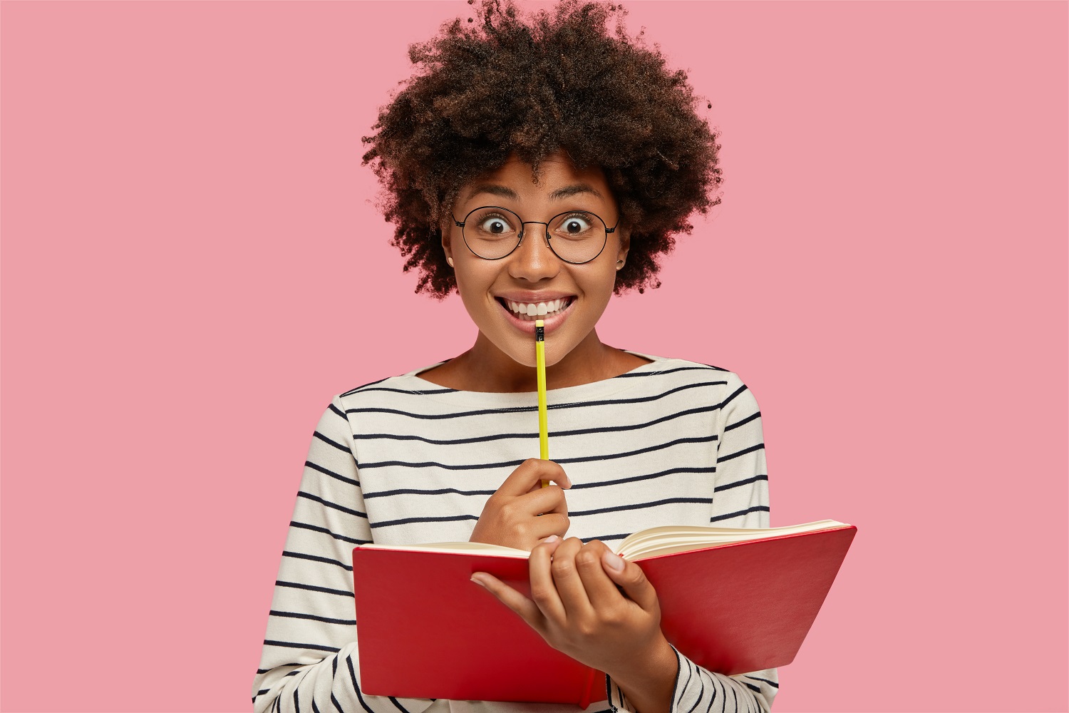 Fundo rosa e mulher sorrindo com lápis e caderno na mão, anotando dicas de revisão de trabalhos acadêmicos.