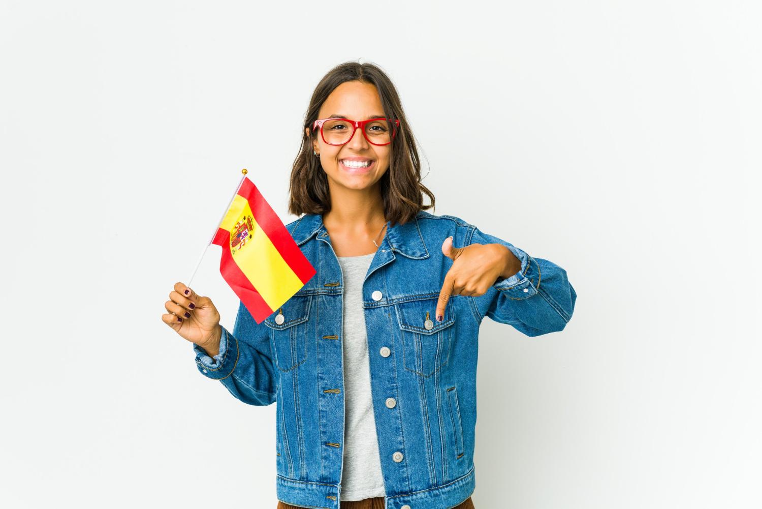 Jovem mulher de óculos segurando bandeira da Espanha, apontando para baixo e sorrindo após descobrir como trabalhar na Espanha.