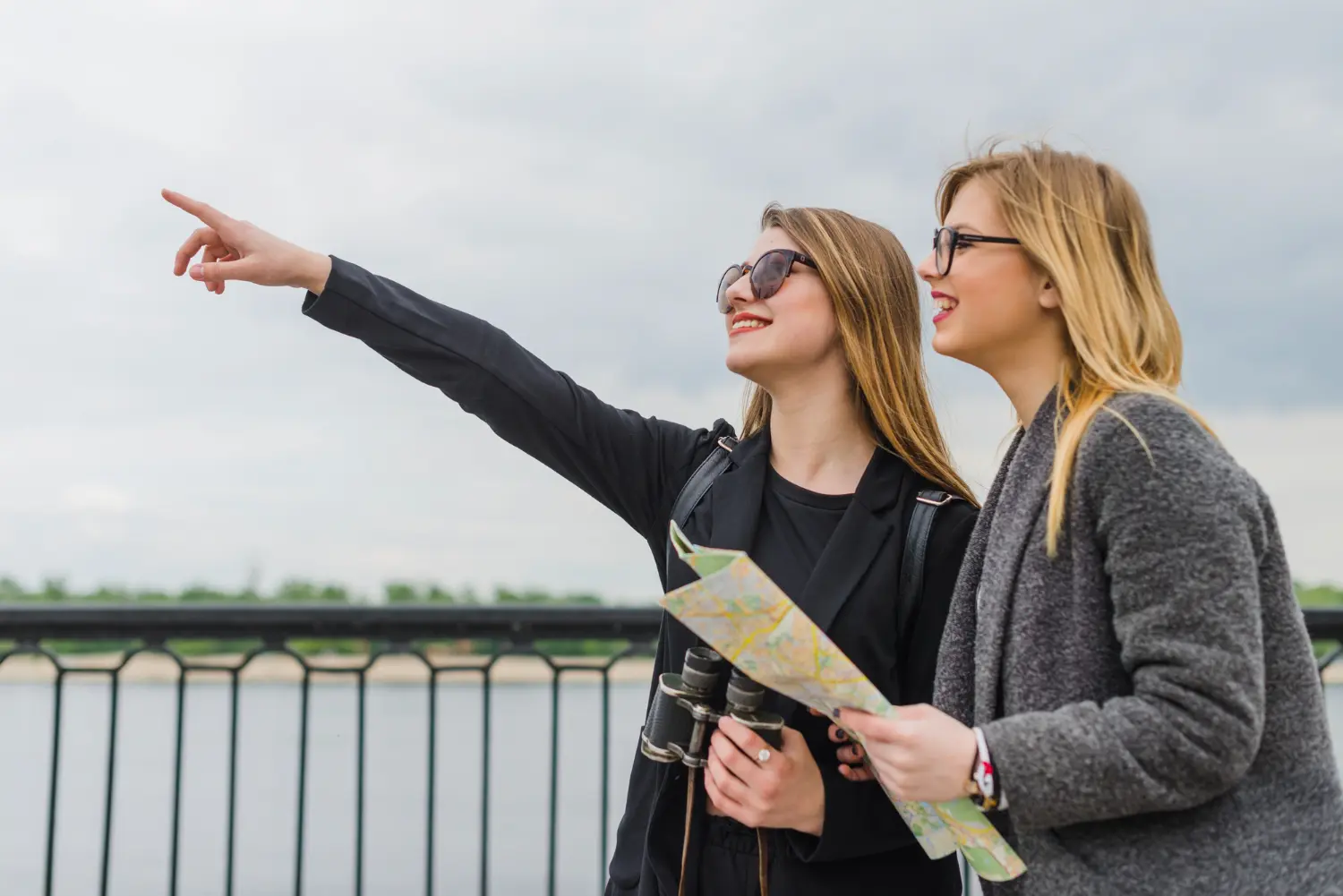 duas amigas turistando, sendo que uma está segurando uma máquina de fotografia e apontando para algum lugar, e a outra, um mapa para saber qual caminho elas vão percorrer