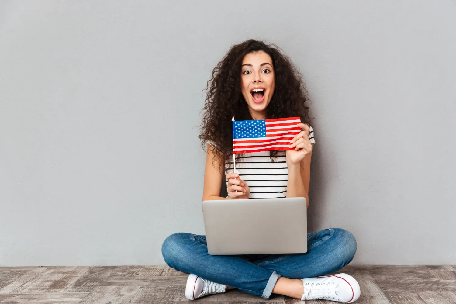 Mulher jovem sentada no chão, encostada numa parede cinza e com as pernas cruzadas, sobre as quais há um notebook, e segurando a bandeira dos EUA